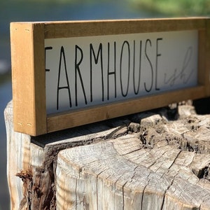 K&ouml;nnte beinhalten: Rechteckiges Holzschild mit den Worten "FARMHOUSE ish" in schwarzer Schrift. Das Schild hat einen hellbraunen Rahmen und wird auf einem Baumstumpf pr&auml;sentiert. Der Hintergrund ist verschwommen, was auf eine Umgebung im Freien hindeutet.