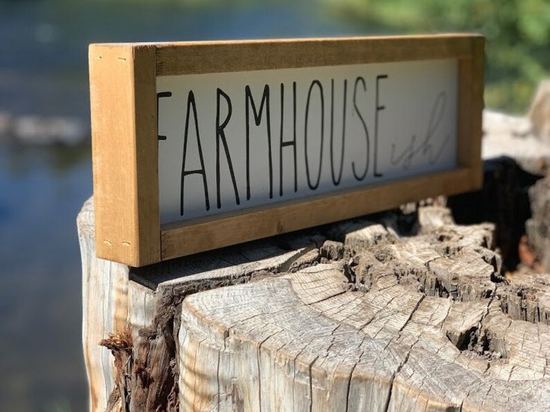 K&ouml;nnte beinhalten: Rechteckiges Holzschild mit den Worten "FARMHOUSE ish" in schwarzer Schrift. Das Schild hat einen hellbraunen Rahmen und wird auf einem Baumstumpf pr&auml;sentiert. Der Hintergrund ist verschwommen, was auf eine Umgebung im Freien hindeutet.