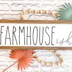 K&ouml;nnte beinhalten: Ein rechteckiges Holzschild mit den Worten "FARMHOUSE ish" in schwarzer Schrift. Das Schild ist in einem dunkelbraunen Holzrahmen eingefasst. Dekorative Holzperlen und getrocknete Palmbl&auml;tter sind um das Schild angeordnet.