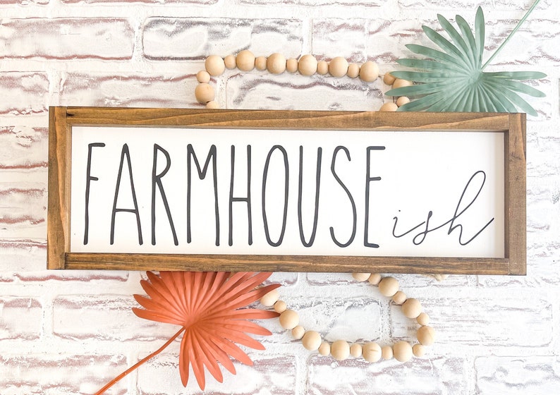 K&ouml;nnte beinhalten: Ein rechteckiges Holzschild mit den Worten "FARMHOUSE ish" in schwarzer Schrift. Das Schild ist in einem dunkelbraunen Holzrahmen eingefasst. Dekorative Holzperlen und getrocknete Palmbl&auml;tter sind um das Schild angeordnet.