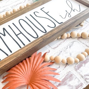 K&ouml;nnte beinhalten: Rechteckiges Holzschild mit dem Schriftzug "FARMHOUSE ish" in Schwarz. Eine Kette aus Holzperlen und eine korallenfarbene Blattdekoration sind ebenfalls zu sehen.