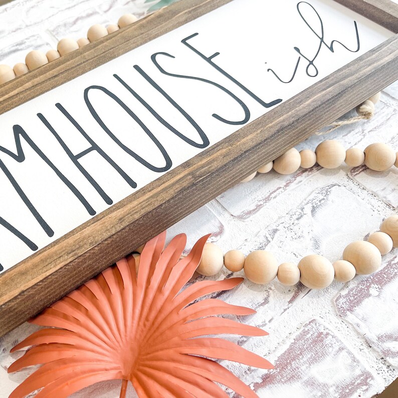 K&ouml;nnte beinhalten: Rechteckiges Holzschild mit dem Schriftzug "FARMHOUSE ish" in Schwarz. Eine Kette aus Holzperlen und eine korallenfarbene Blattdekoration sind ebenfalls zu sehen.