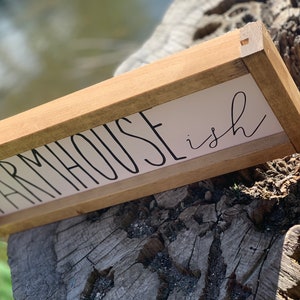 K&ouml;nnte beinhalten: Ein rechteckiges Holzschild mit den Worten "FARMHOUSE ish" in schwarzer Schrift auf wei&szlig;em Hintergrund. Das Schild ist mit hellbraunem Holz umrahmt und liegt auf einem verwitterten Baumstumpf.
