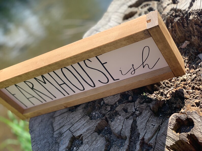 K&ouml;nnte beinhalten: Ein rechteckiges Holzschild mit den Worten "FARMHOUSE ish" in schwarzer Schrift auf wei&szlig;em Hintergrund. Das Schild ist mit hellbraunem Holz umrahmt und liegt auf einem verwitterten Baumstumpf.
