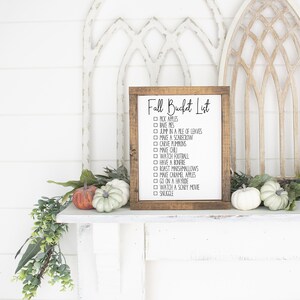 May include: A wooden sign with a brown frame and white background. The sign says "Fall Bucket List" and has a checklist of fall activities, including "pick apples", "make pies", "jump in a pile of leaves", "make a scarecrow", "carve pumpkins", "have a bonfire", "watch football", "roast marshmallows", "make caramel apples", "go on a hayride", "watch a scary movie", and "snuggle".