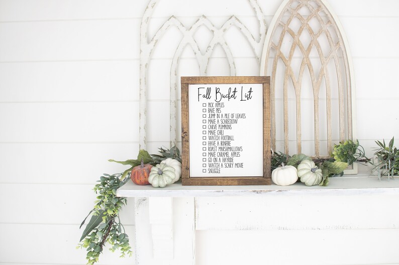 May include: A wooden sign with a brown frame and white background. The sign says "Fall Bucket List" and has a checklist of fall activities, including "pick apples", "make pies", "jump in a pile of leaves", "make a scarecrow", "carve pumpkins", "have a bonfire", "watch football", "roast marshmallows", "make caramel apples", "go on a hayride", "watch a scary movie", and "snuggle".