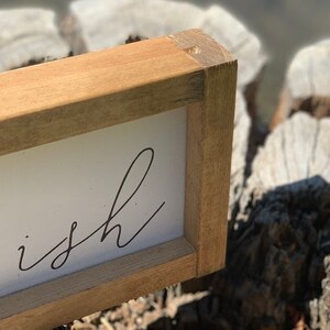 K&ouml;nnte beinhalten: Rechteckiges Holzschild mit dem Wort "ish" in schwarzer Schrift auf wei&szlig;em Hintergrund. Der Rahmen besteht aus hellbraunem Holz. Das Schild ist handgefertigt und hat eine rustikale &Auml;sthetik.