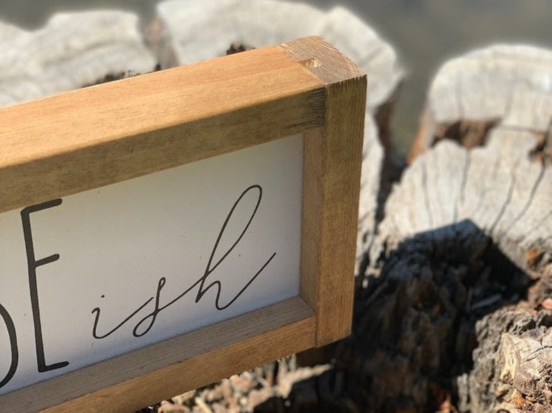 K&ouml;nnte beinhalten: Rechteckiges Holzschild mit dem Wort "ish" in schwarzer Schrift auf wei&szlig;em Hintergrund. Der Rahmen besteht aus hellbraunem Holz. Das Schild ist handgefertigt und hat eine rustikale &Auml;sthetik.