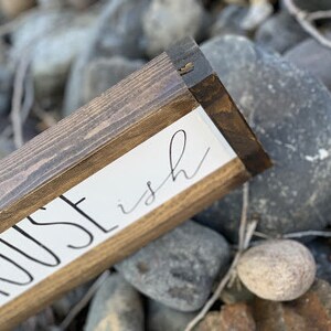 K&ouml;nnte beinhalten: Holzschild mit den Worten "HOUSE ish" in schwarzer Schrift auf wei&szlig;em Hintergrund. Das Schild hat einen dunkelbraunen Holzrahmen und liegt auf einem Bett aus grauen Steinen.