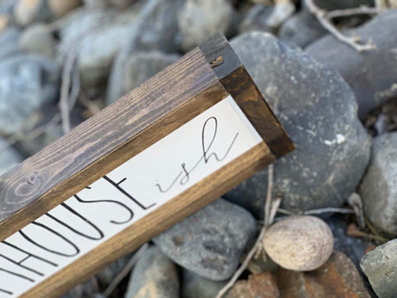K&ouml;nnte beinhalten: Holzschild mit den Worten "HOUSE ish" in schwarzer Schrift auf wei&szlig;em Hintergrund. Das Schild hat einen dunkelbraunen Holzrahmen und liegt auf einem Bett aus grauen Steinen.