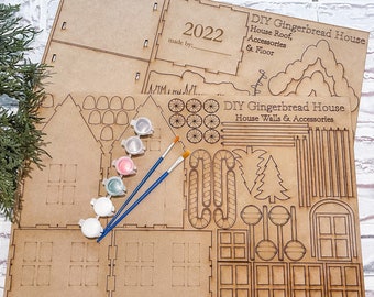 Può includere: Un kit fai-da-te per costruire una casetta di pan di zenzero con pezzi di legno per le pareti, il tetto e gli accessori. Il kit include barattoli di vernice, pennelli e istruzioni. Il kit è etichettato "DIY Gingerbread House" e "2022".