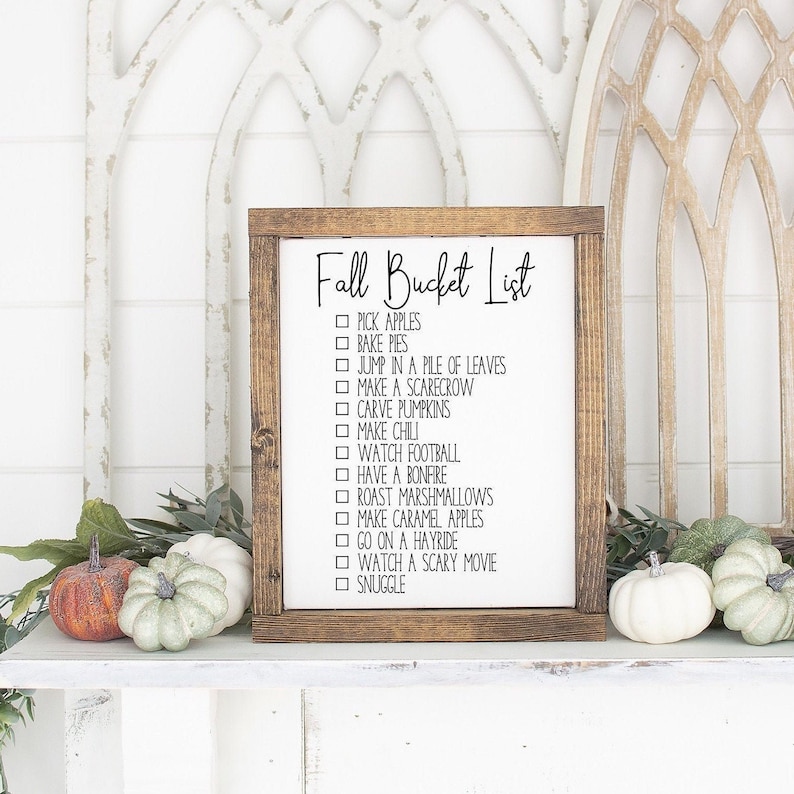 May include: A wooden sign with the text "Fall Bucket List" and a checklist of fall activities, including picking apples, baking pies, jumping in a pile of leaves, making a scarecrow, carving pumpkins, making chili, watching football, having a bonfire, roasting marshmallows, making caramel apples, going on a hayride, watching a scary movie, and snuggling.