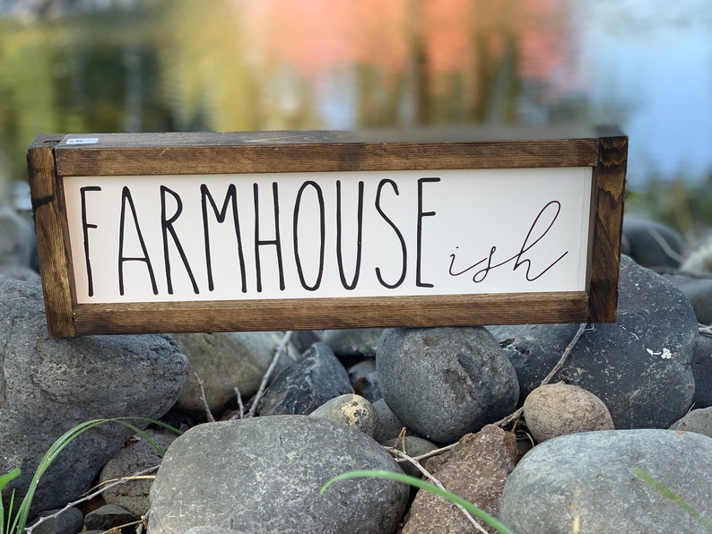 K&ouml;nnte beinhalten: Ein rechteckiges Holzschild mit den Worten "FARMHOUSE ish" in schwarzer Schrift auf wei&szlig;em Hintergrund. Das Schild hat einen dunkelbraunen Rahmen und liegt auf grauen Steinen.