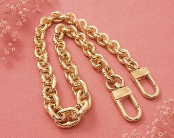 Puede incluir: Una correa de cadena de metal en tono dorado con eslabones gruesos y redondeados entrelazados. La cadena termina en dos mosquetones a juego en cada extremo, diseñados para sujetarse a bolsos o accesorios con una superficie pulida.