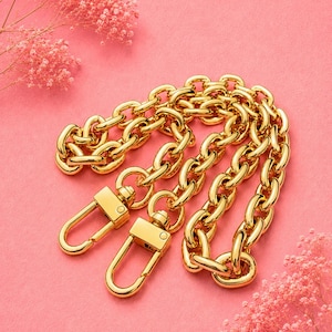 May include: A gold-coloured chain with two clasp closures. The chain is made of large, rounded links. The background is a solid pink colour, with pink flowers in the corners.