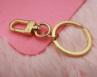 May include: Gold-toned metal key chain with a swivel clasp and a round ring. The key chain is on a pink fuzzy surface.