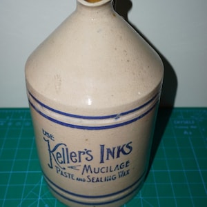Op de afbeelding: Een beige stenen kruik met een handvat en een smalle tuit. De kruik heeft blauwe horizontale strepen en de blauwe tekst: USE Keller's INKS MUCILAGE PASTE AND SEALING WAX. Het object staat op een groene snijmat met zichtbare maataanduidingen.
