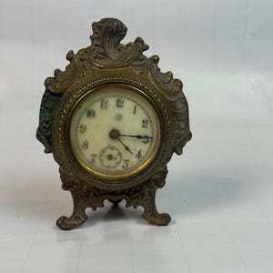 May include: A small, ornate brass-toned metal table clock featuring a circular off-white face with black Arabic numerals and a secondary sub-dial. The clock is encased in a decorative, scrolled metal frame with three feet and a top finial.