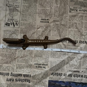 May include: A brown metal alligator figurine positioned above a white measuring tape that shows the object is approximately 8 inches long. The alligator has a textured back, a long tail, and four legs, with its mouth slightly open.