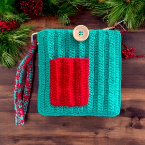 Puede incluir: Un bolso de crochet turquesa con un bolsillo de crochet rojo y un botón blanco. El bolso tiene una larga correa de crochet turquesa y roja.