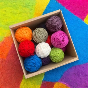 May include: A box filled with colorful yarn balls in various shades of red, blue, green, orange, and white. The yarn balls are arranged in a circular pattern, with the largest ball in the center.