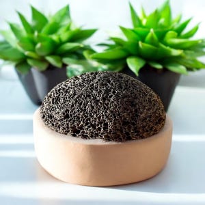 Op de afbeelding: Een zwarte vulkanische puimsteen in een beige ronde container. De steen is ruw en getextureerd. De container is gemaakt van een zacht materiaal.