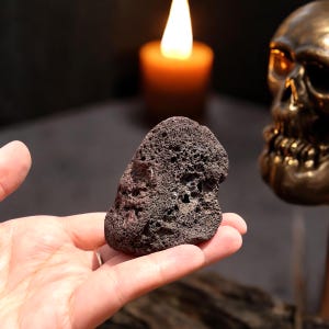 Peut inclure: Une roche de couleur gris foncé, rugueuse et poreuse avec une surface texturée. La roche est tenue dans une main.