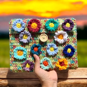 Könnte beinhalten: Eine grüne gehäkelte Clutch mit einer Vielzahl von bunten gehäkelten Blumen, darunter rote, gelbe, blaue, weiße und lila Blumen. Die Clutch hat einen Verschluss aus Holz.