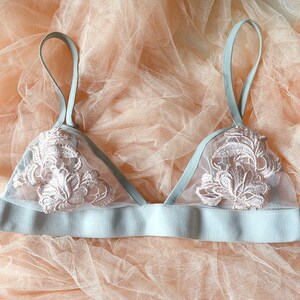 Könnte beinhalten: Hellblaues Bralette mit zarter rosa-weißer Blumenstickerei. Es hat ein geripptes Band und dünne Träger. Der Hintergrund ist ein durchsichtiges, pfirsichfarbenes Gewebe. Dessous.