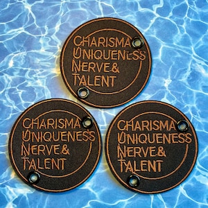 May include: Three black circular patches with the words "CHARISMA, UNIQUENESS, NERVE & TALENT" embroidered in a copper color. Each patch has a copper-colored border and three black eyelets. The patches are on a blue water-like background.