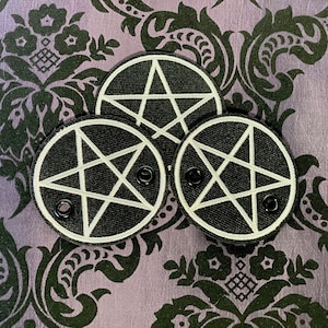 May include: Three black and white pentagram patches. Each patch features a white embroidered pentagram within a black circle. The patches have black snap fasteners and are set against a purple background with a black floral pattern.