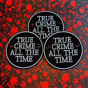 May include: Three black circular patches with white embroidered text reading "TRUE CRIME ALL THE TIME." Each patch has a white border and two eyelets. The background is a red and black splatter pattern.