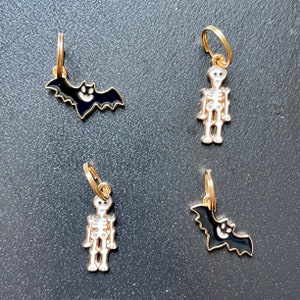 May include: Four Halloween-themed charms. Two charms are black bats with gold trim and a smiling face. The other two charms are white skeletons with gold trim. Each charm has a gold hoop for hanging.