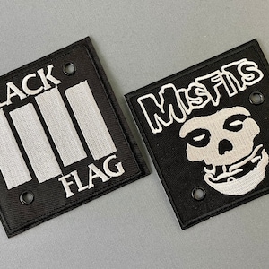 Puede incluir: Dos parches cuadrados de tela negra. Uno presenta el texto "BLACK FLAG" con tres barras verticales blancas. El otro parche muestra la palabra "MISFITS" sobre un gráfico de calavera blanca. Ambos parches tienen detalles bordados en blanco.