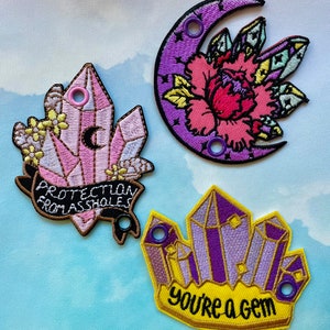 May include: Embroidered patches featuring crystal, moon, and floral designs. One patch reads "PROTECTION FROM ASSHOLES," another features a crescent moon and flower, and the third says "you're a gem."