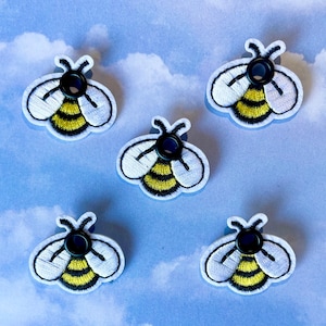 May include: Five embroidered bee-shaped patches. Each patch features a white body with black outline, yellow and black striped abdomen, and a black button center. The patches are arranged on a light blue background with a cloud pattern.