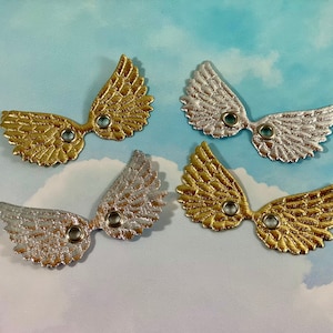 May include: Four metallic wing-shaped embellishments in gold and silver. Each wing has a detailed feather design and a small hole for attachment. The wings are arranged on a sky-blue background.