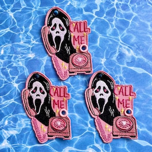 May include: Embroidered patches featuring the Ghostface mask from the Scream movies, holding a pink telephone. The patches have the words "CALL ME" in pink, with a pink phone and a black background. The patches are on a blue background.