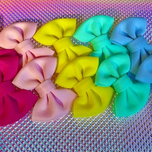 May include: A collection of colorful fabric bows in shades of pink, yellow, green, and blue. The bows are arranged on a shimmering, iridescent surface, creating a vibrant and eye-catching display. These bows are suitable for hair accessories or craft projects.