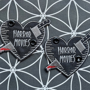 May include: Two black heart-shaped patches with the words "HORROR MOVIES" embroidered in white. Each patch features a silver cleaver and a bloody knife. The patches have black grommets.