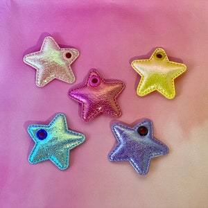 May include: Five star-shaped ornaments in iridescent colors: pink, purple, blue, and yellow. Each star has a small hole at the top for hanging. The ornaments are made of a shiny, textured material and are arranged on a pink and purple gradient background.