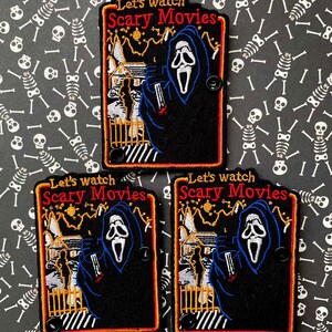 May include: Embroidered patches with the text "Let's watch Scary Movies" in red and yellow. The patches feature a black hooded figure with a white mask, holding a knife, in front of a house and fence. The background is black.