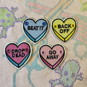 May include: Four heart-shaped patches with embroidered text and eyelets. The patches are in light blue, yellow, pink, and pink with the phrases "BEAT IT", "BACK OFF", "DROP DEAD", and "GO AWAY". Each patch has a white border and a black outline.