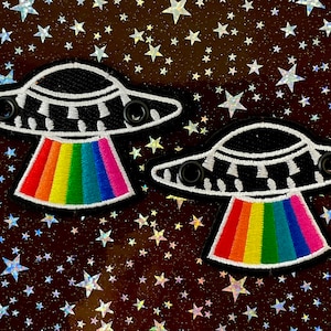 May include: Two embroidered patches featuring a black and white UFO design with a rainbow beam. The patches have a black border and are set against a starry background. Each UFO has two black circular details.