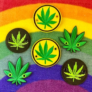 May include: A collection of embroidered patches and appliqués featuring cannabis leaf designs. The patches are in various colors, including yellow, black, and green, with black borders and eye-shaped details. The background is a rainbow-colored fabric.