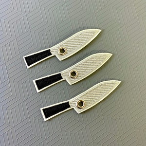 May include: Three embroidered knife patches with silver blades, black handles, and a silver grommet near the handle. The patches are arranged on a gray surface with a geometric pattern.