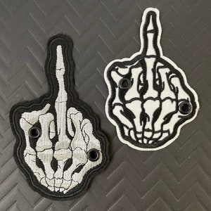 May include: Two embroidered patches in the shape of a hand giving the middle finger. One patch has a black border with a gray skeleton hand design. The other has a white background with a black skeleton hand design.
