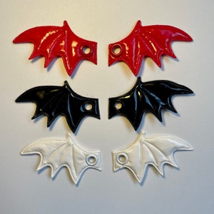 May include: Three pairs of bat wing-shaped accessories in red, black, and white. Each wing has a glossy finish and a circular hole for attachment. The wings are arranged in a symmetrical pattern, showcasing their design and color variations.