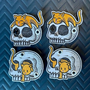 May include: Four embroidered patches featuring a skull design with a cat. Two patches show a cat resting on top of the skull, while the other two have a cat emerging from the skull. The patches are white, black, and orange.