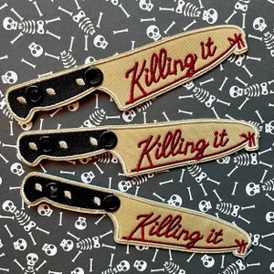 May include: Three embroidered patches in the shape of a knife with the words "Killing it" in burgundy script. The knife blades are tan, and the handles are black with three black buttons. The background is black with white skeleton and bone designs.
